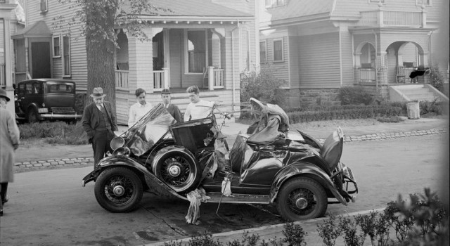 Giving a rare glimpse of the day's fashion, a group of men look over a crumpled car that sits by the side of a residential Boston street