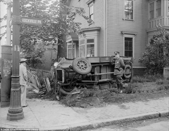 A police officer poses next to a car that flipped over maneuvering around corner in Roxbury, Massachusetts in 1935.